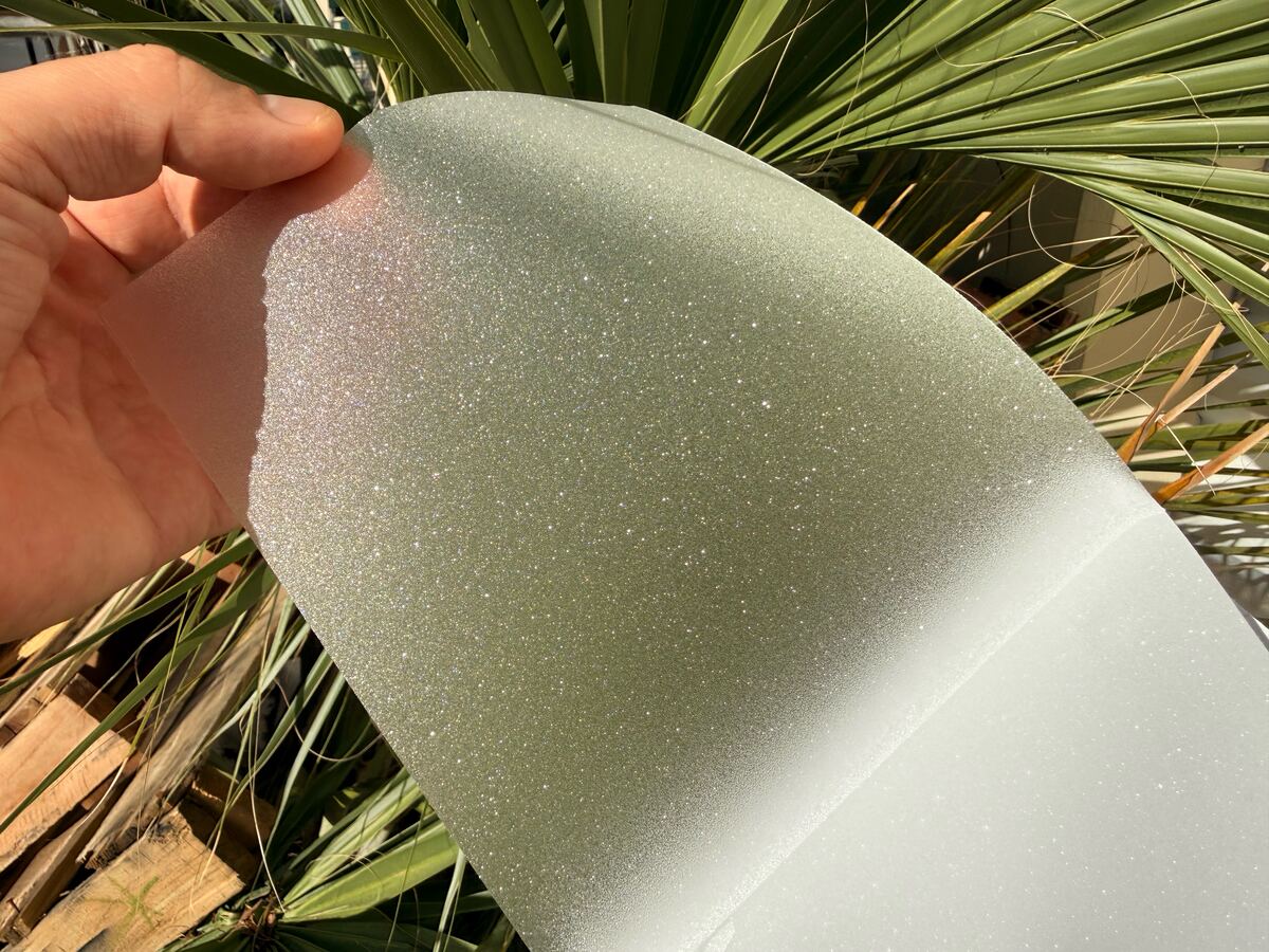 A sheet of SHINECOR frosted film, partly peeled from its white liner, held up in bright sun: the crystal grain sparkles.